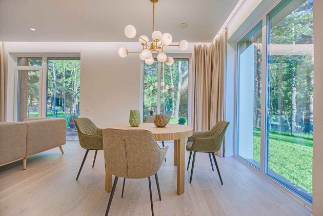Bright dining area with a round wood table, upholstered chairs, and a modern globe chandelier beside floor-to-ceiling windows.