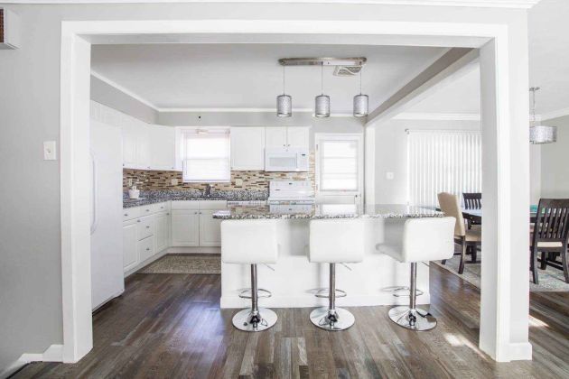 Bright, open-concept kitchen with white cabinetry, mosaic backsplash, and a granite peninsula lined with four white swivel barstools; pendant lights hang overhead and a dining area with vertical blinds is visible to the right.