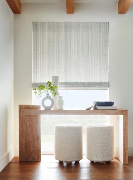 Bright entry nook featuring a light gray, subtly striped woven shade fully lowered over a tall window; a chunky wooden console holds white ceramic decor and greenery, with two plush boucle stools tucked underneath.
