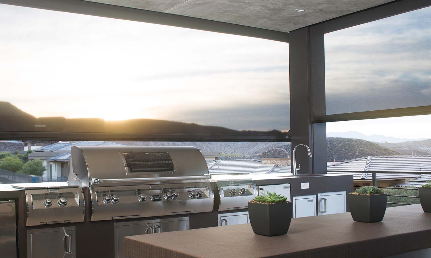 Covered outdoor kitchen with stainless gas grills and a sink, framed by large motorized solar screens that filter the setting sun and reveal mountain and rooftop views; three small succulent planters sit on the counter.