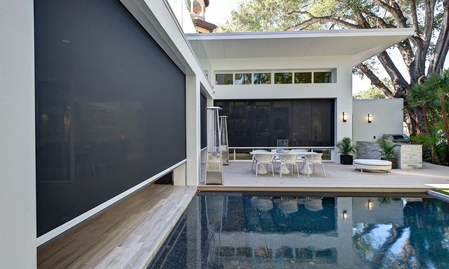 Modern backyard with a rectangular swimming pool beside a white contemporary house, featuring large dark retractable screens over the glass walls and a raised wooden deck with a dining table, heaters, and outdoor seating under tall trees.
