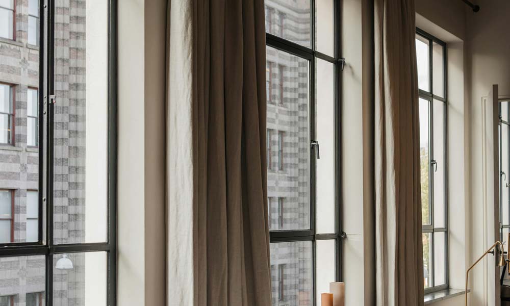 Close-up of tall steel-frame windows dressed in heavy, neutral drapery panels, softening exterior city sounds while adding warmth to the interior.