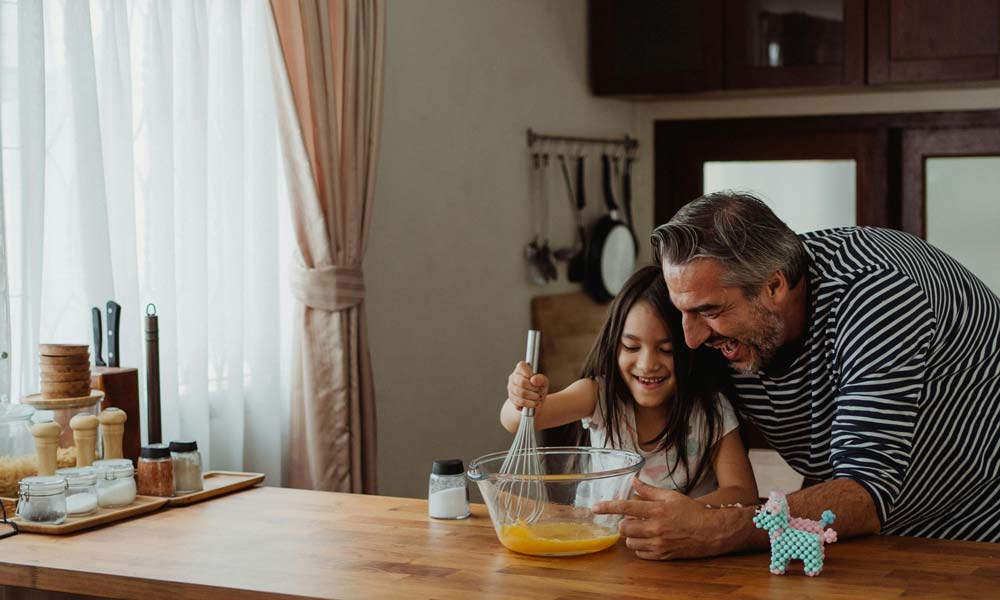 Smiling man and young girl whisking eggs together in a glass bowl at a wooden kitchen island, with sunlight filtering through curtains and cooking utensils and spice jars in the background.