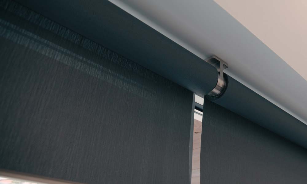 Tight close-up of two dark fabric roller shades with exposed cylindrical tubes and metal mounting brackets, showing the texture of the material and the smooth, minimalist hardware along the ceiling.