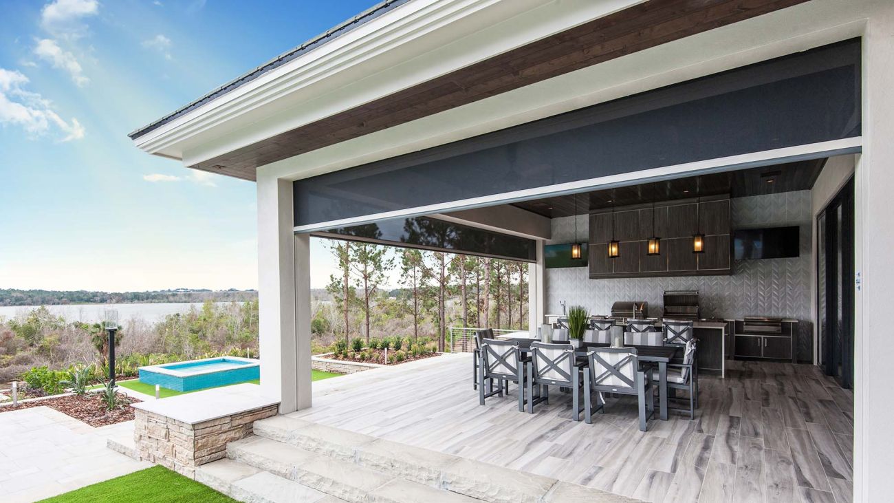 Covered outdoor kitchen and dining area with a large retractable screen partially lowered, overlooking a pool and scenic view.