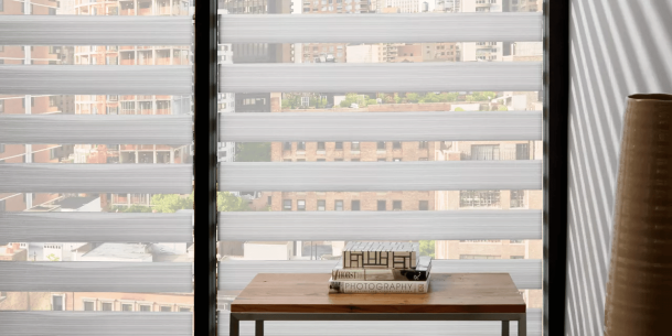 Modern banded shades with alternating sheer and opaque horizontal stripes filtering daylight across a high-rise cityscape; small console table with stacked design books in the foreground.