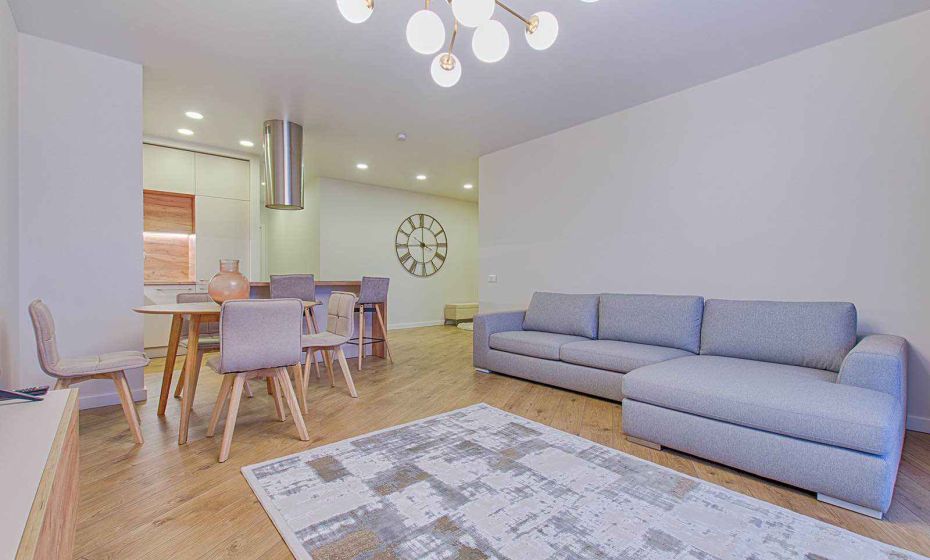 Modern living room with a gray sectional sofa, wooden dining table with upholstered chairs, large wall clock, and contemporary chandelier in a bright open-concept home.